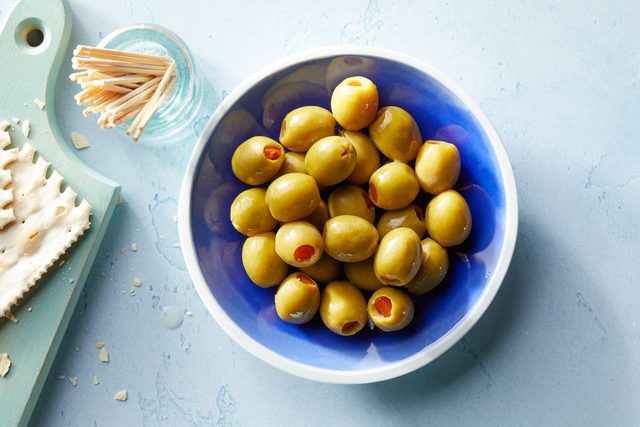 Manzanilla olives in a blue bowl on blue background, glass jar of toothpicks nearby