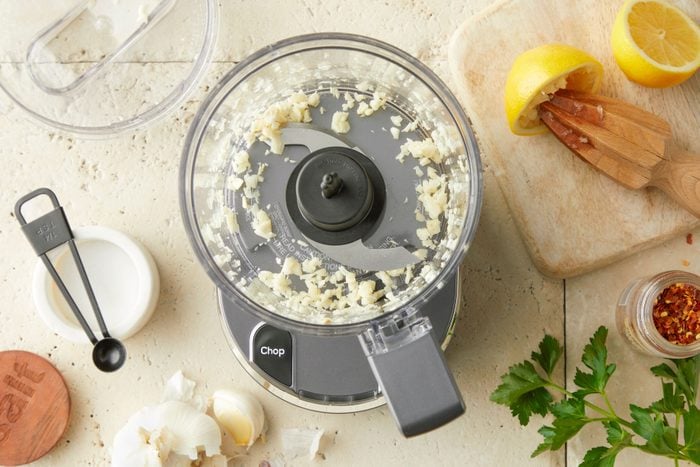 Minced garlic in a blender with lemon and red chilli placed on the side on a marble countertop