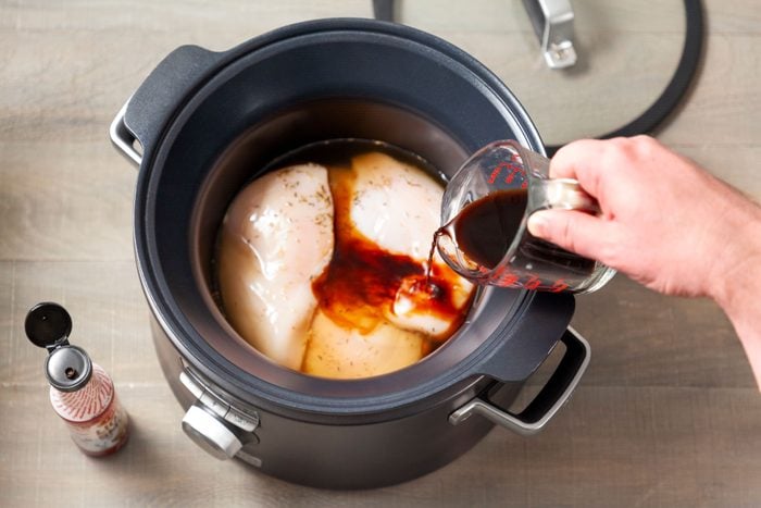 Pouring liquid smoke over raw chicken in slow cooker on wooden surface