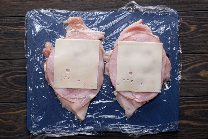 slices of ham and cheese placed over flattened chicken breasts on a cutting board on wooden surface