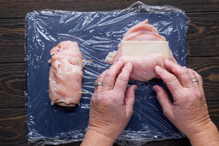 A Person's Hands Rolling Chicken Breast with Cheese Slice on a Cutting Board on Wooden Surface