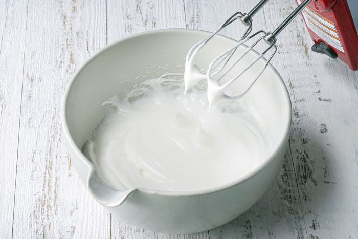Beating Egg Whites With A Hand Mixer In A Large White Bowl on White Painted Wooden Surface