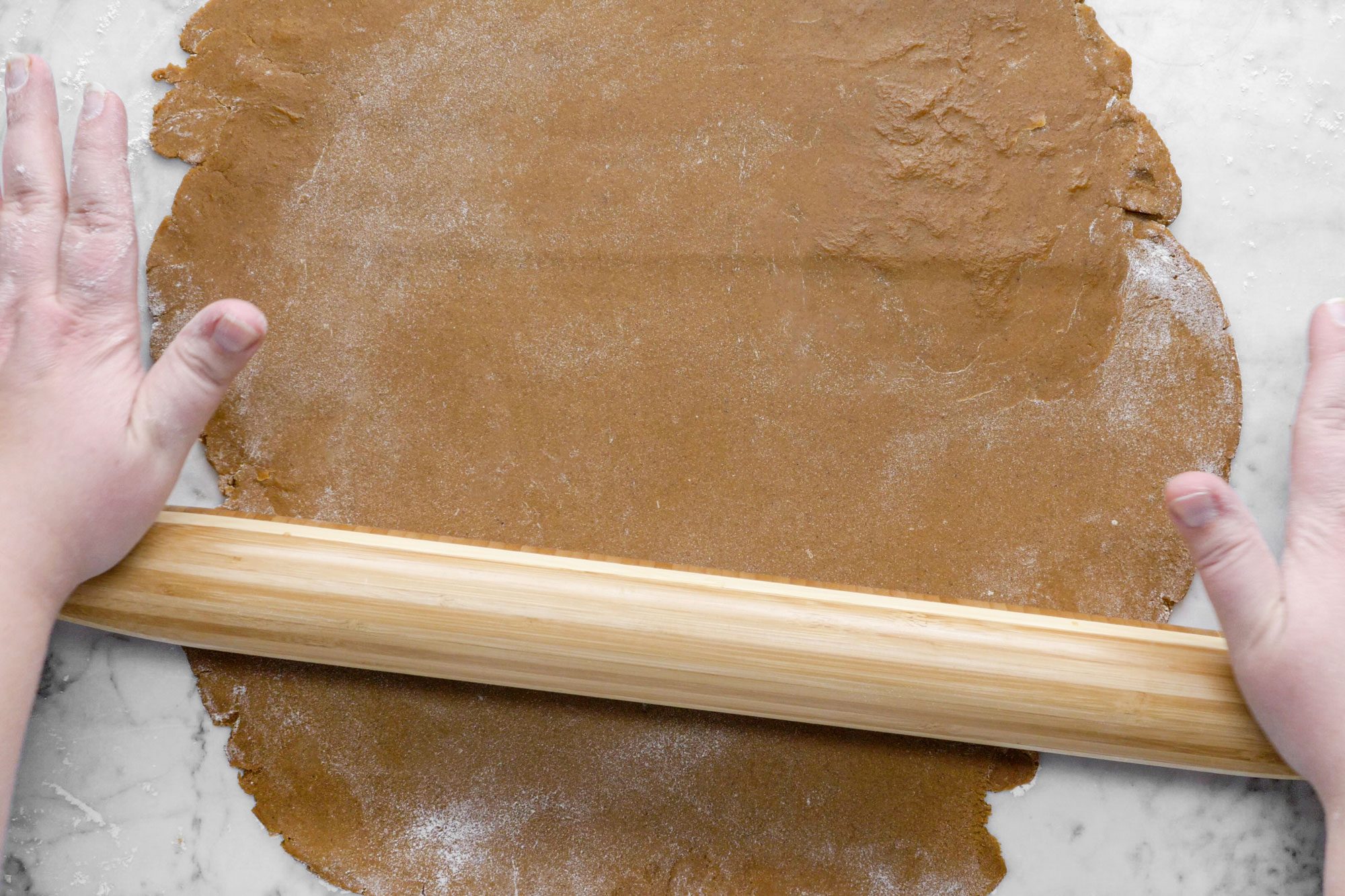 A Woman Rolling Gingerbread Cookie Dough with Wooden Rolling Pin with Hands on Marble Surface