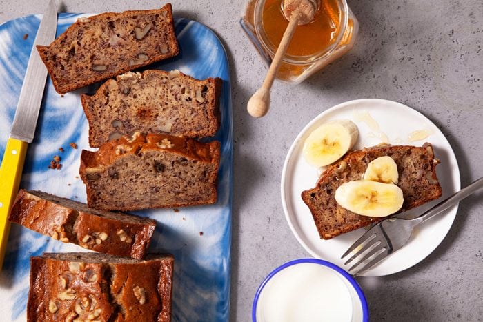 Gluten-Free Banana Bread with a Knife on a Fancy Chopping Board and Pieces of Gluten Free Banana Bread Served on a Small Plate with Banana Slices Next to It.