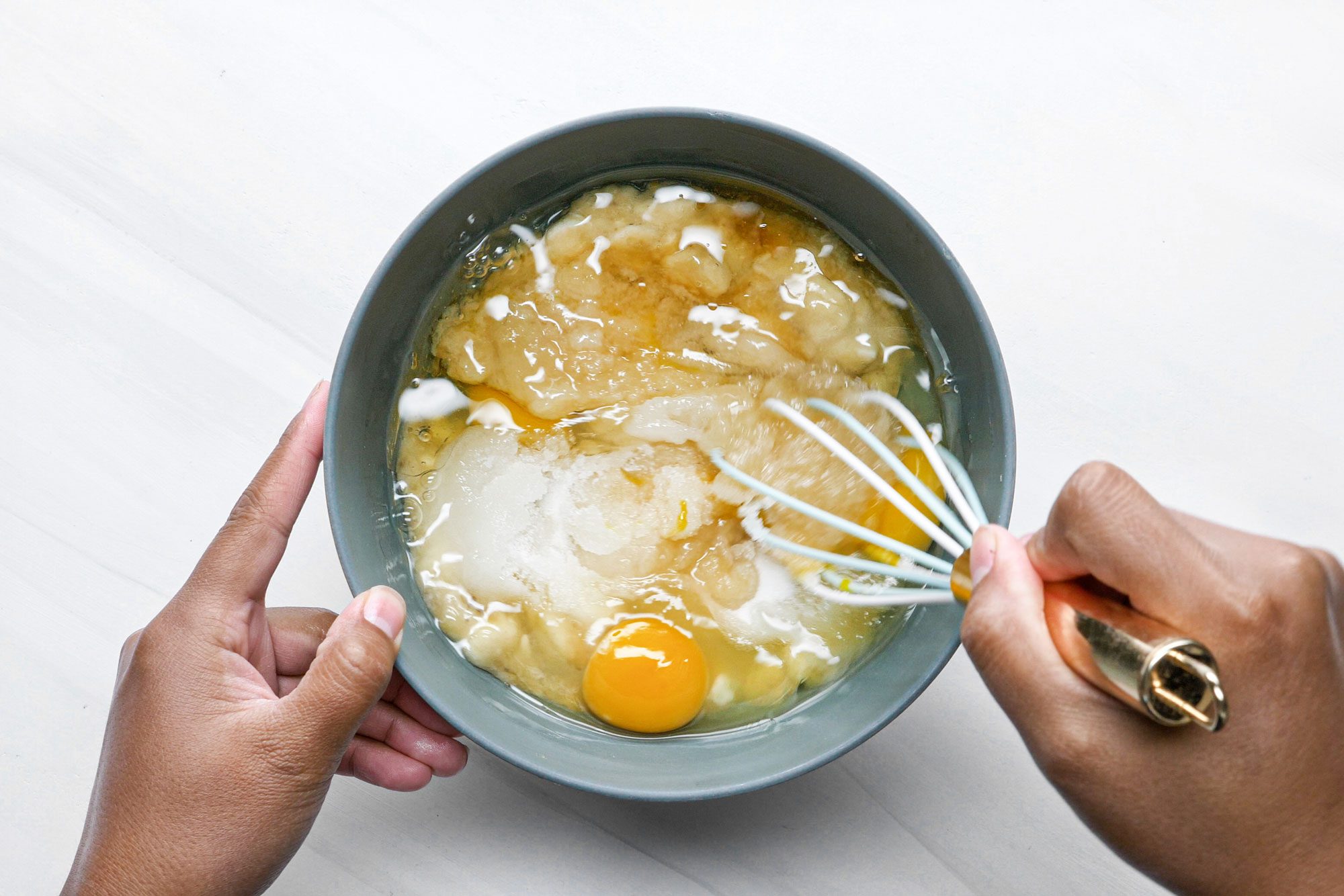A person mixing eggs, bananas, sugar, applesauce, oil, and vanilla in a large bowl using a whisk