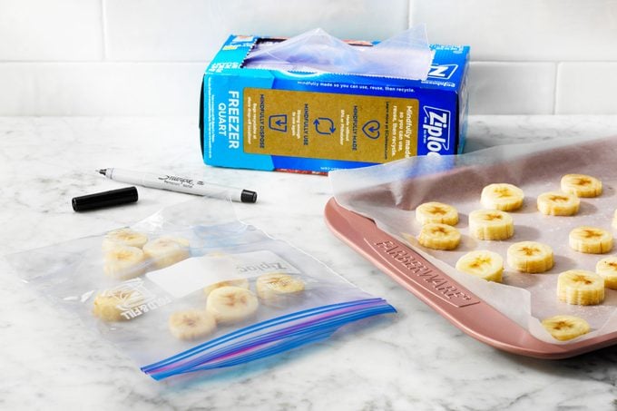 On a kitchen counter, frozen slices of banana in a freezer bag is next to a baking sheet of frozen banana slices