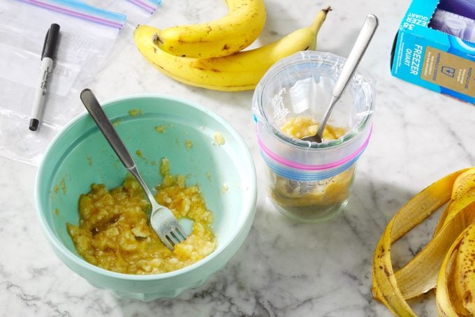 Mashed banana being spooned from a bowl into a freezer bag being held open with a mason jar. Extra freezer bags, permanent marker, banana peels, and whole bananas are nearby on the counter