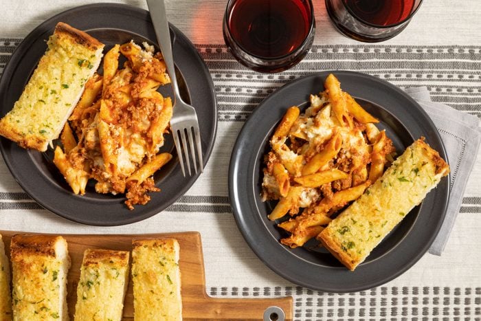 Mostaccioli pasta and bread in a casserole dish on a table with wine served on the side