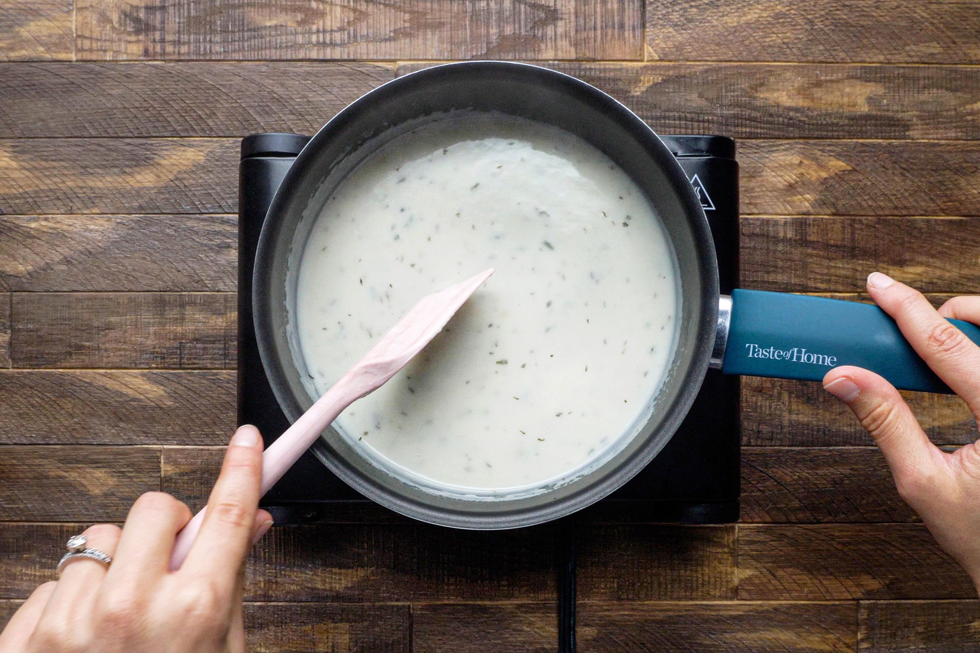 hands stirring the flour, dried parsley flakes, salt, dried thyme, pepper and milk together in a saucepan on a gas stove on a wooden surface