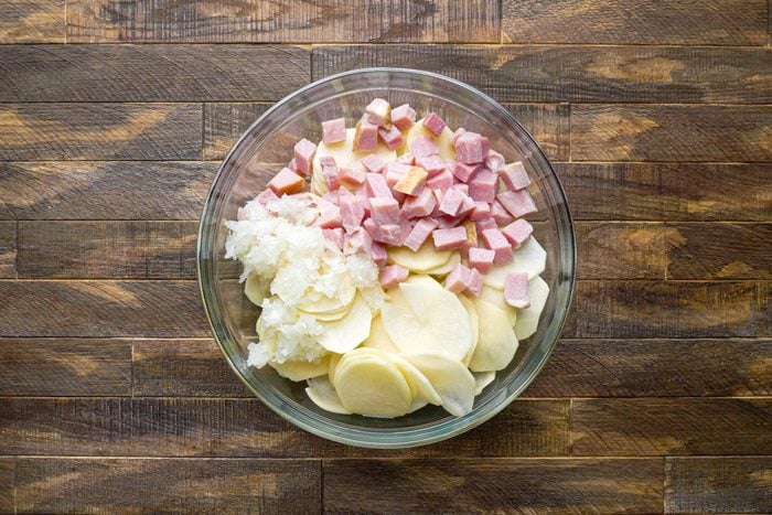 sliced potatoes, chopped ham and grated onion in a large glass bowl on a wooden surface