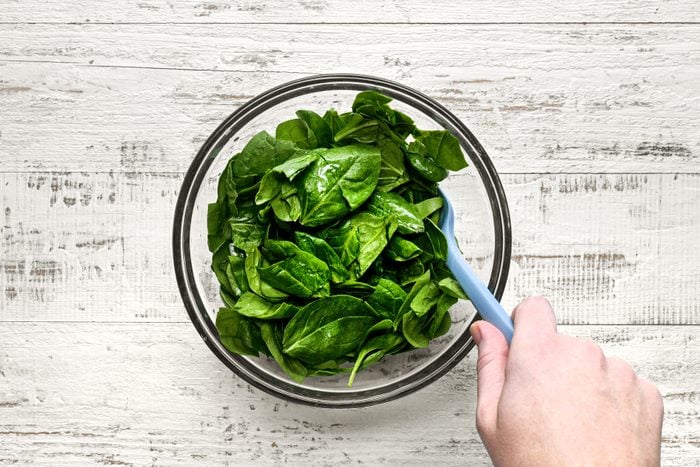 A hand with a blue spoon over a glass bowl of spinach.