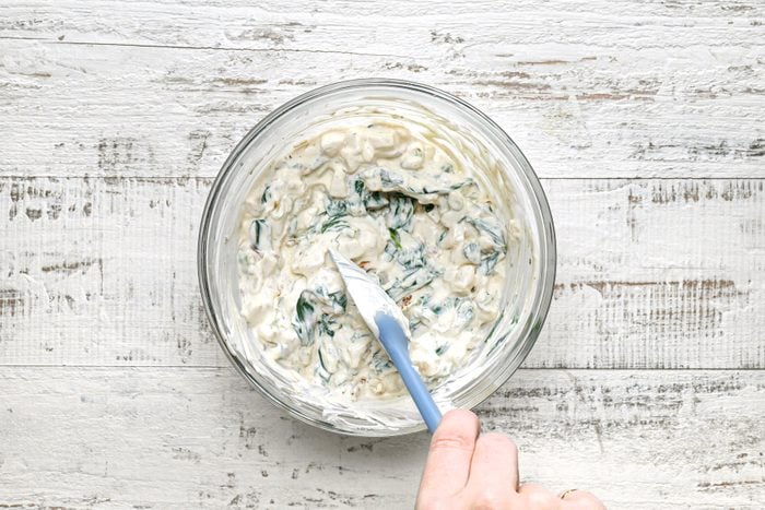 A hand holding a blue spoon over a bowl of spinach dip mixture.