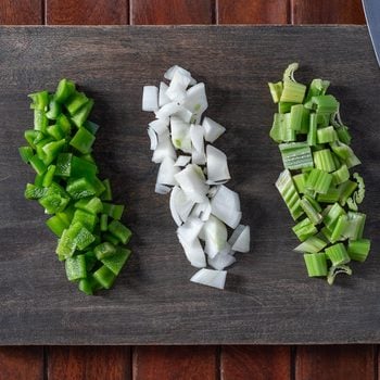 Chopped Green Bell Peppers, white onions and Celery on a wooden chopping board with knife