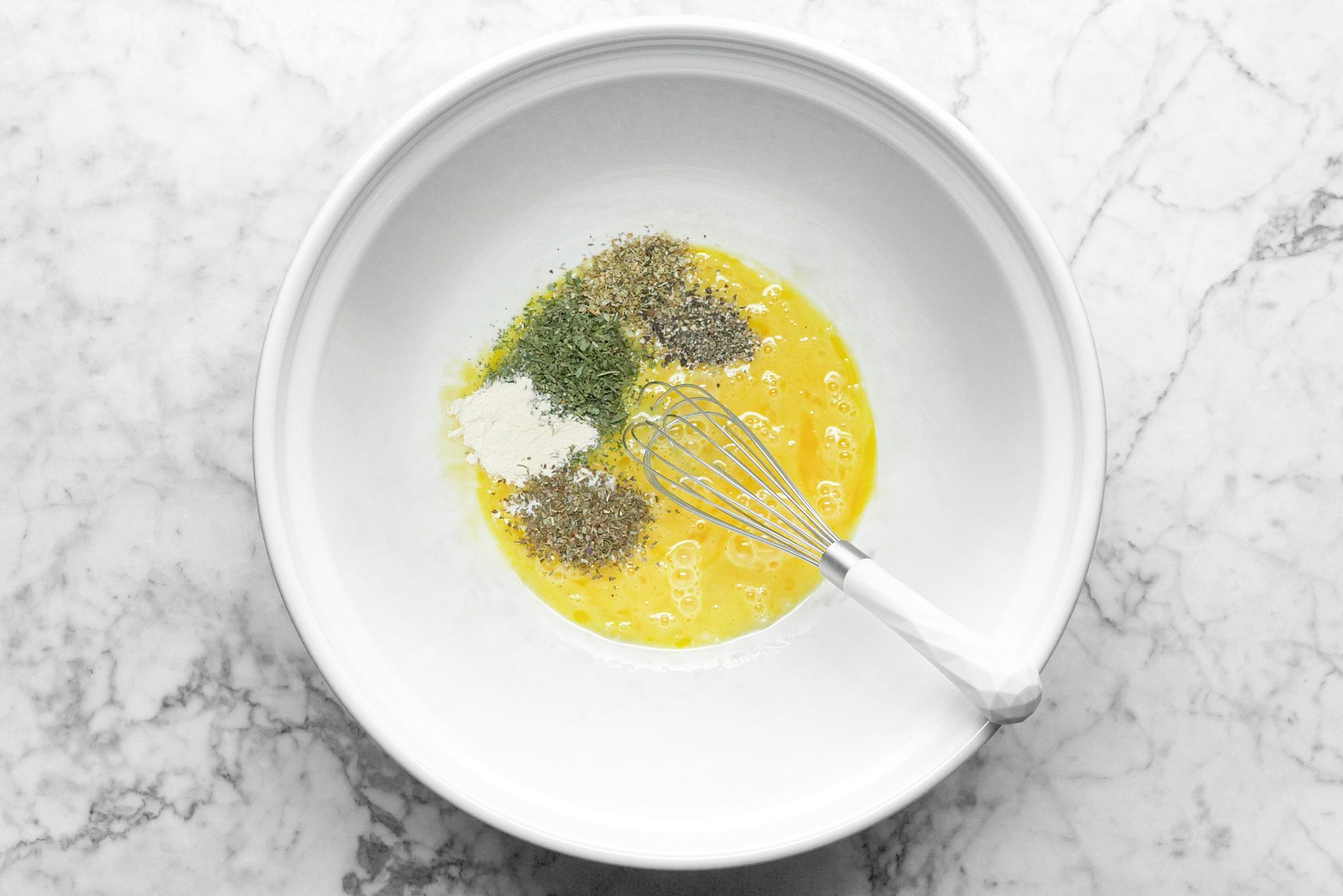 egg, parsley, salt, garlic powder, oregano and pepper in a large white ceramic bowl with a whisk on marble surface