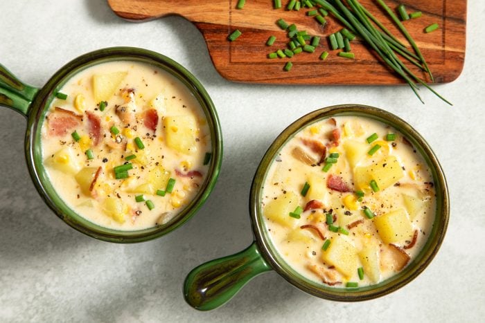 Two bowls of corn chowder with bacon soup next to a cutting board