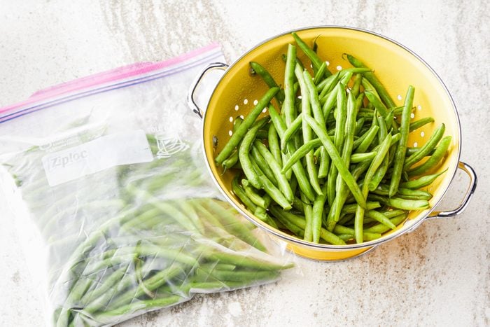 overhead view of frozen green beans in a plastic freezer bag and a colander of fresh green beans on a marble counter top surface
