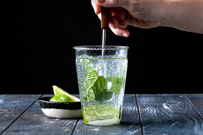 a hand stirring Mojito using a spoon in a highball glass