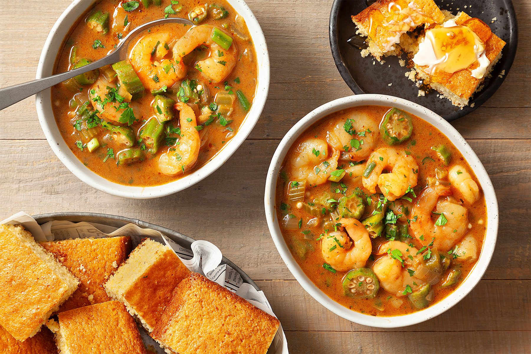 overhead shot; wooden background; Seafood Gumbo; served in two white bowls; with spoon; breads; in serving basket; one bread piece; in black small plate;