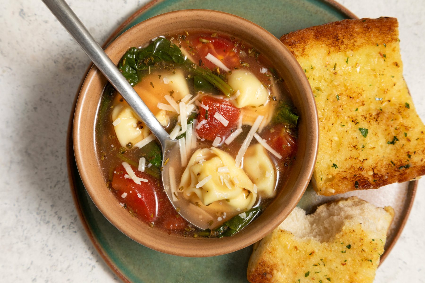 A bowl of spinach tortellini soup with a spoon next to a piece of bread