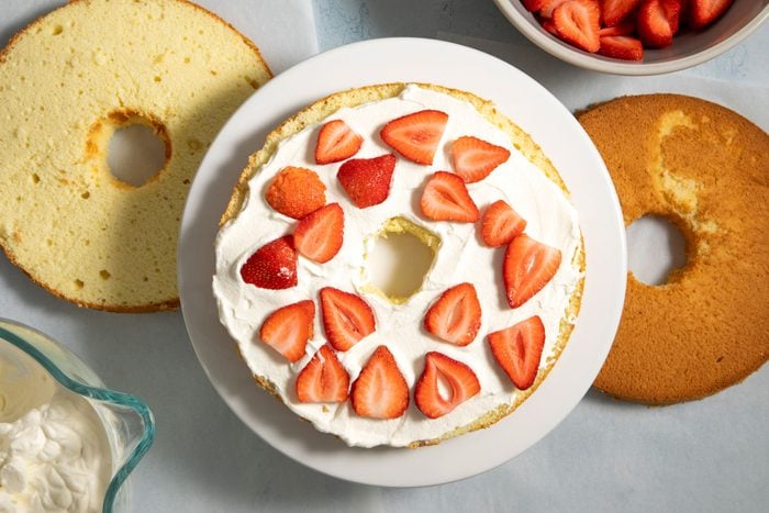 Placing one layer of cake on a serving plate with whipped cream and sliced strawberries