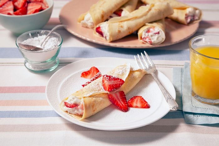 A plate of Strawberry Crepes topped with fresh strawberries, served alongside a glass of orange juice