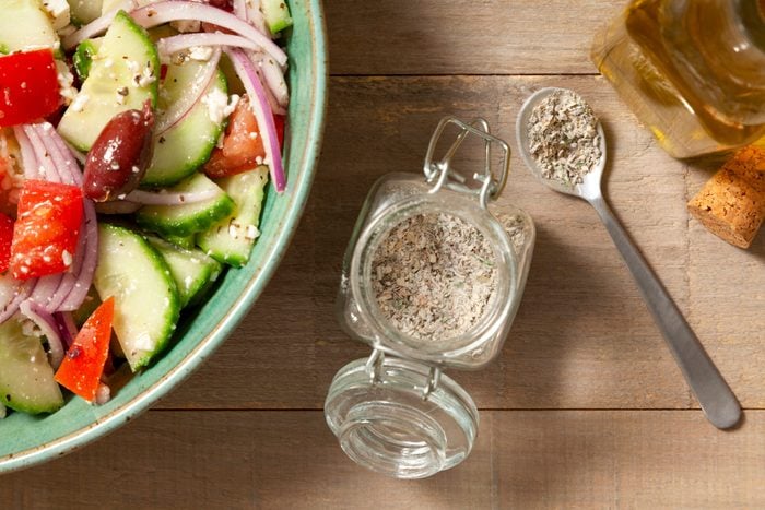Greek Seasoning in a glass air tight jar placed on a wooden surface with a bowl of salad next to it.