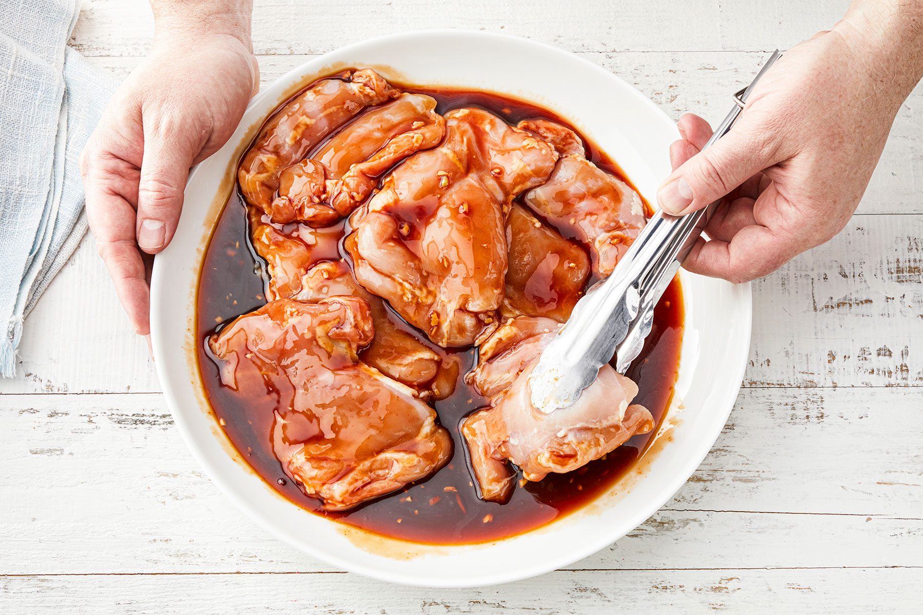 Overhead shot of coat chicken thighs in marinade mixture; tongs; kitchen towel; white wooden background;