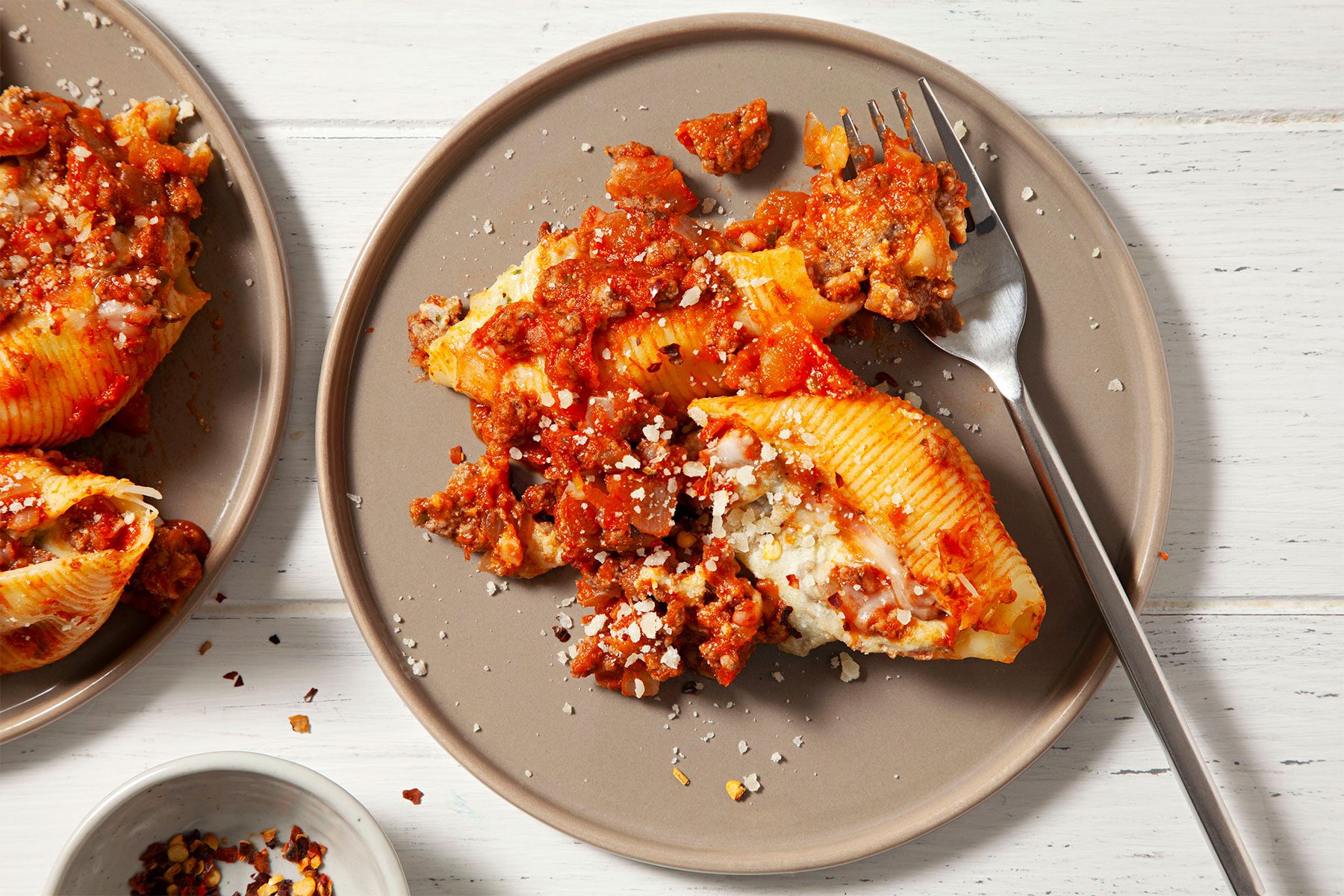 Overhead shot of Easy Beef-Stuffed Shells served in two plates; fork; a small bowl of chilli flakes; white wooden background;