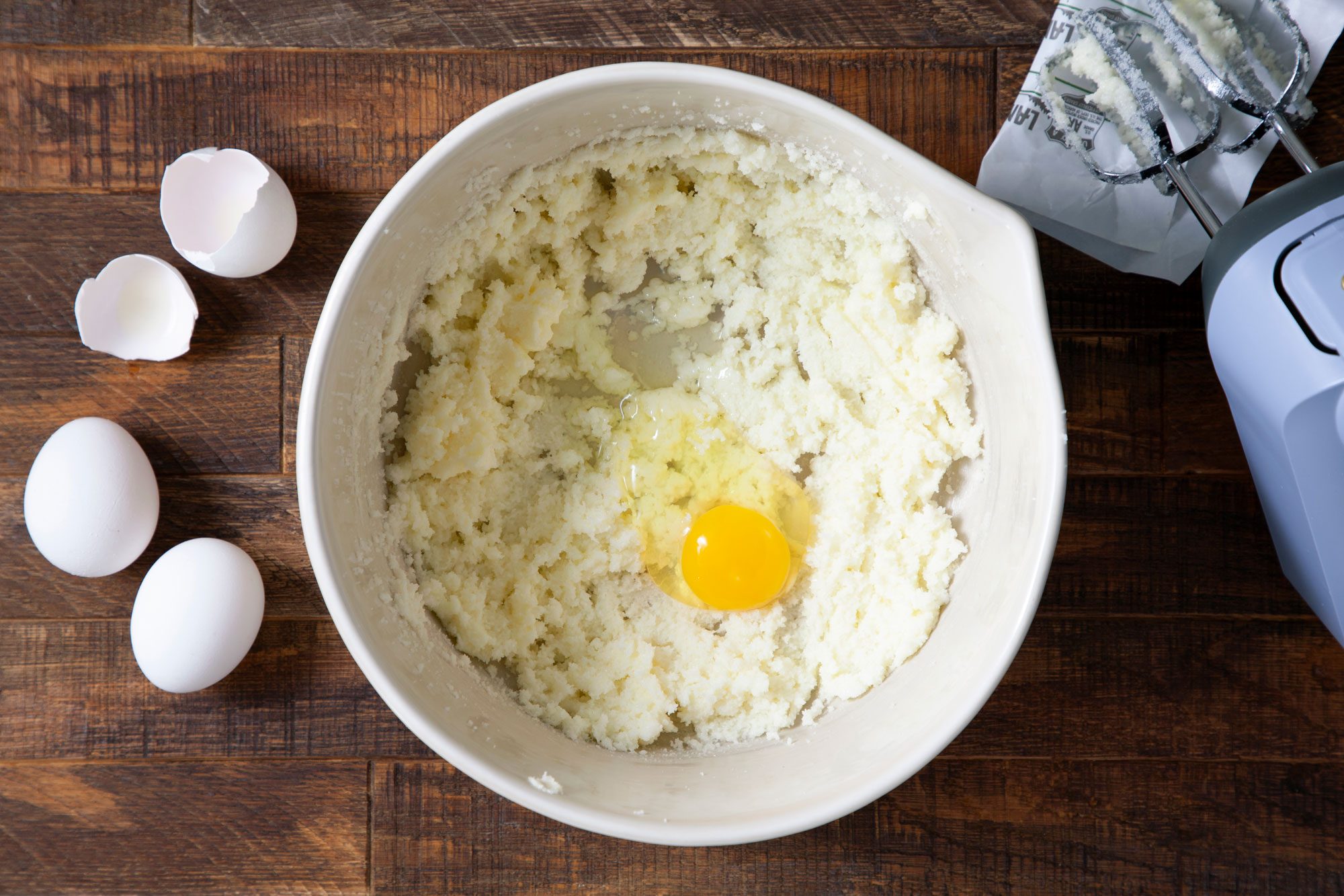 overhead shot of creamed butter and egg in a large bowl and hand mixer on the right on wooden surface