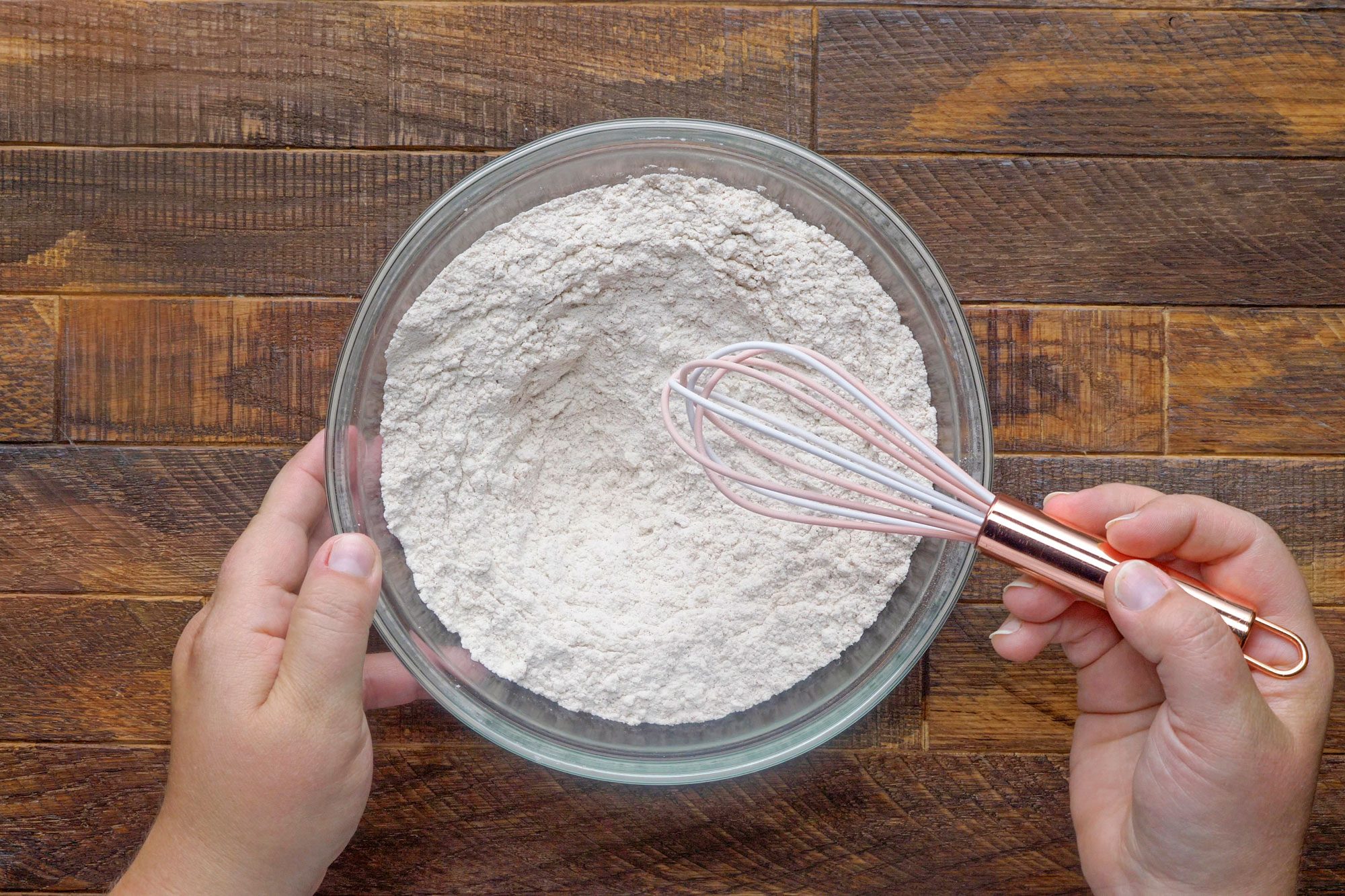 overhead shot of mixing flour, cinnamon, baking soda, salt, and baking powder using whisk in a large glass bowl on wooden surface