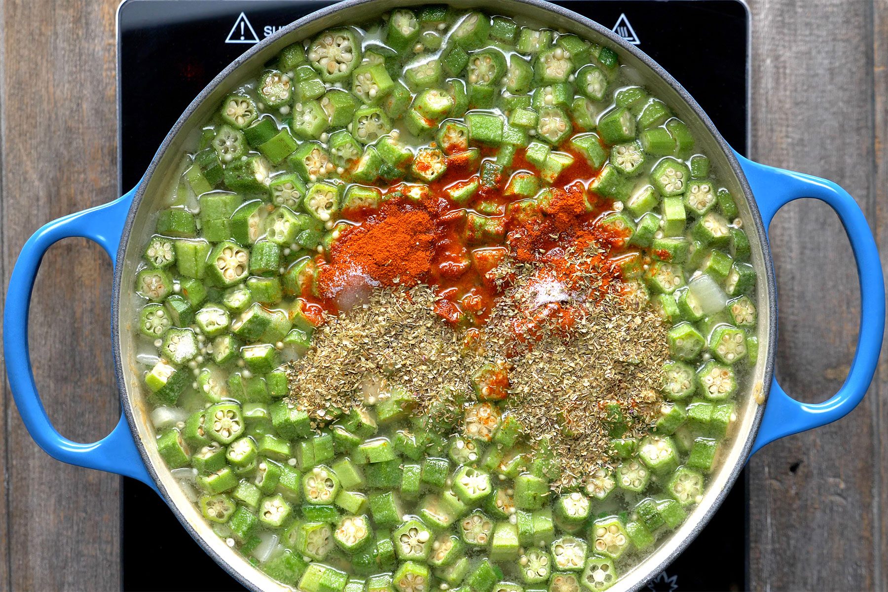 Overhead shot of added the chicken broth; water; okra; paprika; salt; oregano and pepper; bring to boil; wooden background;